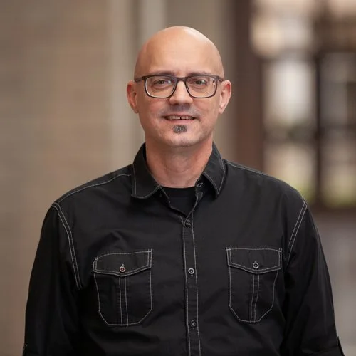 Bald man wearing glasses and a black button-up shirt smiling indoors