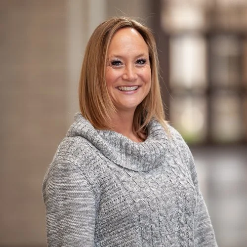 A woman wearing a gray knit sweater smiling in a softly focused indoor setting.