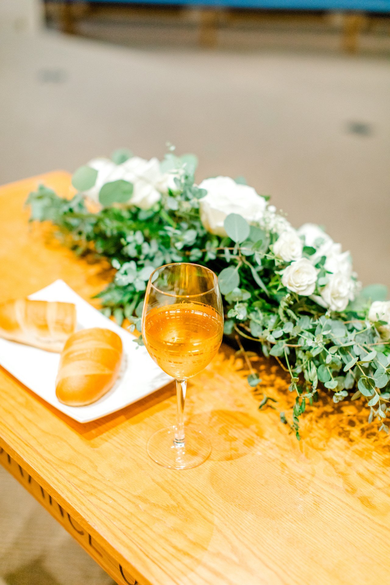 A glass of amber liquid on a wooden table with a plate of bread and floral arrangement.