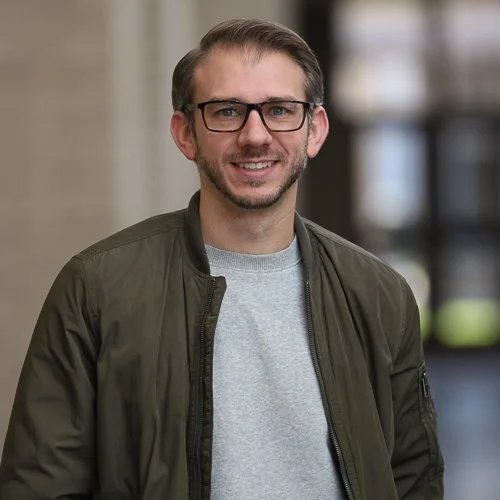 Smiling man with glasses wearing a green jacket and gray shirt indoors.