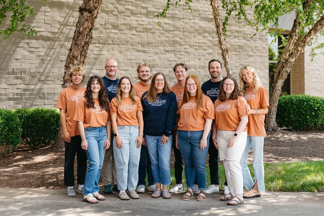 A group of ten young adults posing outdoors in front of a brick wall and trees, some wearing matching orange and navy blue shirts with that read 'Southland Residency'.