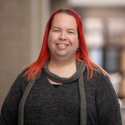 Person with long red hair wearing a gray sweater and striped scarf, smiling in a blurred indoor setting.