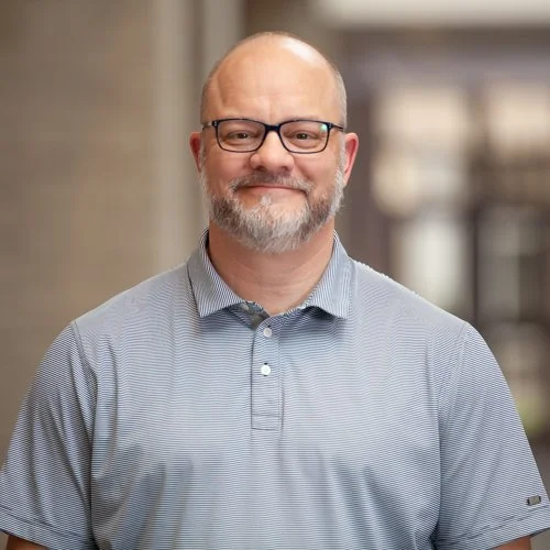 A smiling man with glasses and a beard wearing a gray striped polo shirt indoors.