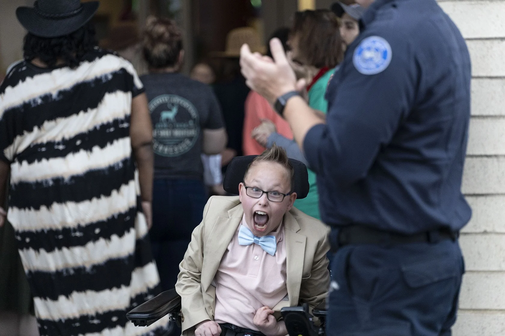 A person in a wheelchair wearing a beige jacket, pink shirt, and blue bowtie, expressing excitement. Background shows a group of people gathered, including a uniformed individual on the right side.