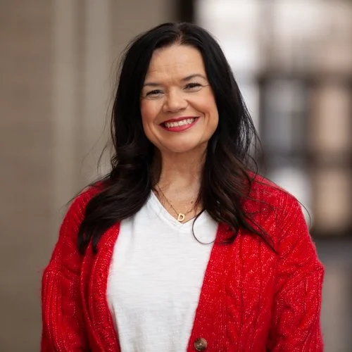 Smiling woman with long dark hair wearing a red cardigan over a white shirt, standing indoors.