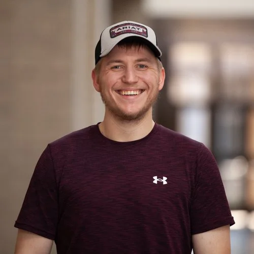Smiling person wearing a dark maroon Under Armour shirt and a baseball cap standing indoors.