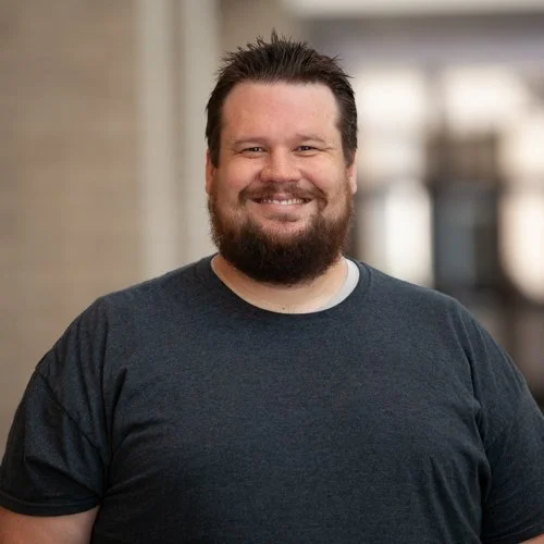 Man with beard smiling, wearing a dark t-shirt in a blurred urban setting.