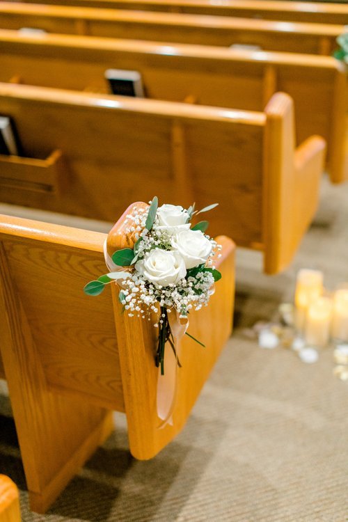 White roses and baby's breath bouquet attached to a wooden church pew, with candles in the background.