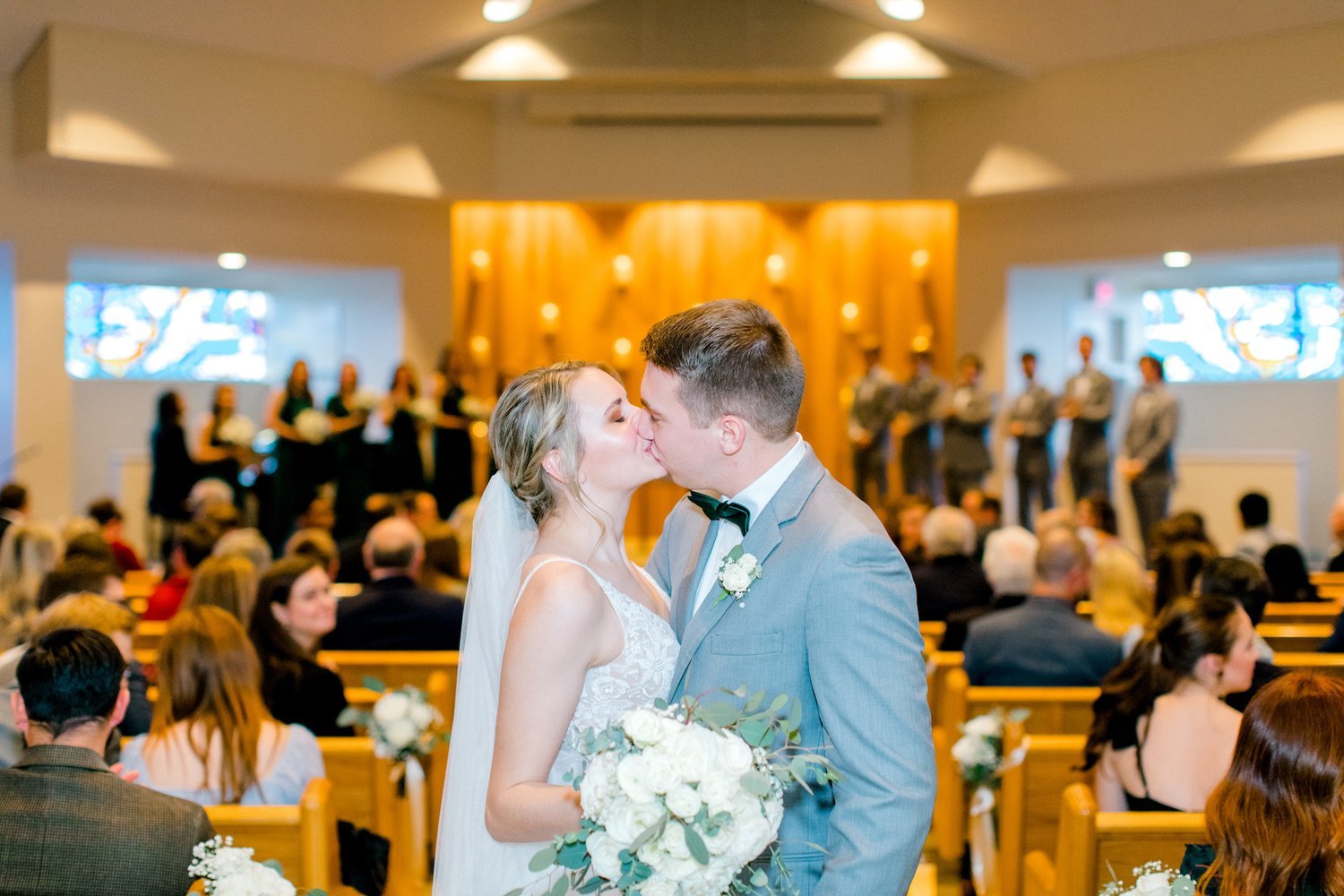 Bride and groom kissing in a church during a wedding ceremony, with bridesmaids and groomsmen in the background.