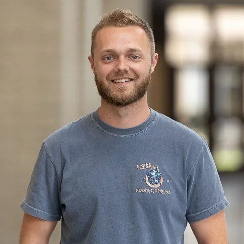 Man with a beard and short hair smiling, wearing a blue T-shirt with "Topsail North Carolina" print, indoors.