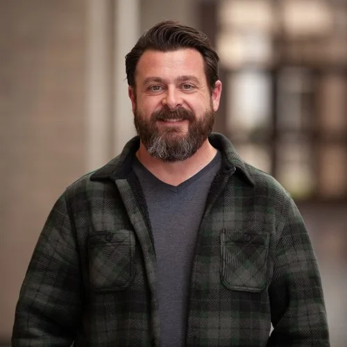 Man with a beard smiling, wearing a green plaid jacket and gray shirt, standing indoors.