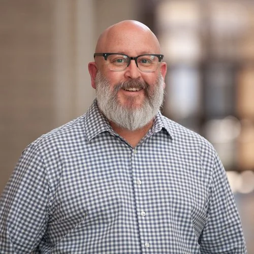 Bald man with beard smiling, wearing glasses and a checkered shirt, blurred background.