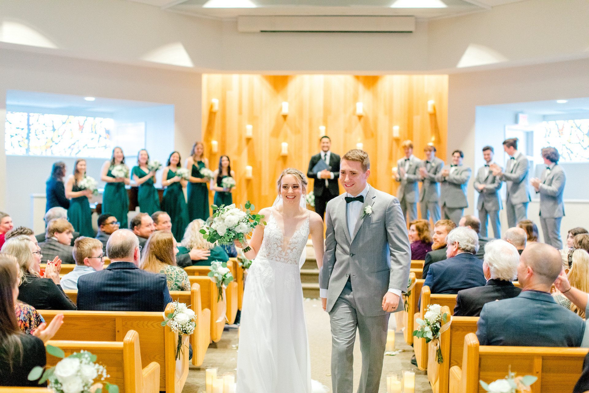 A bride and groom walking down the aisle in a church, surrounded by guests and bridal party. The bride is holding a bouquet, and the groom is wearing a gray suit. Bridesmaids in green dresses and groomsmen in gray suits are visible in the background.