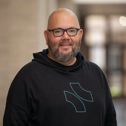 Smiling man with glasses and beard wearing a black hoodie with a logo, standing indoors.