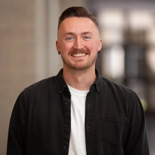 Smiling person wearing a black shirt and white undershirt, standing indoors.