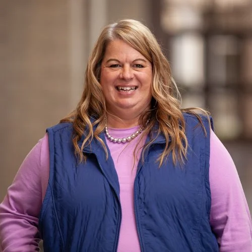 A woman with long blonde hair, wearing a blue vest over a pink shirt, is smiling at the camera. The background is blurred and appears to be outdoors.