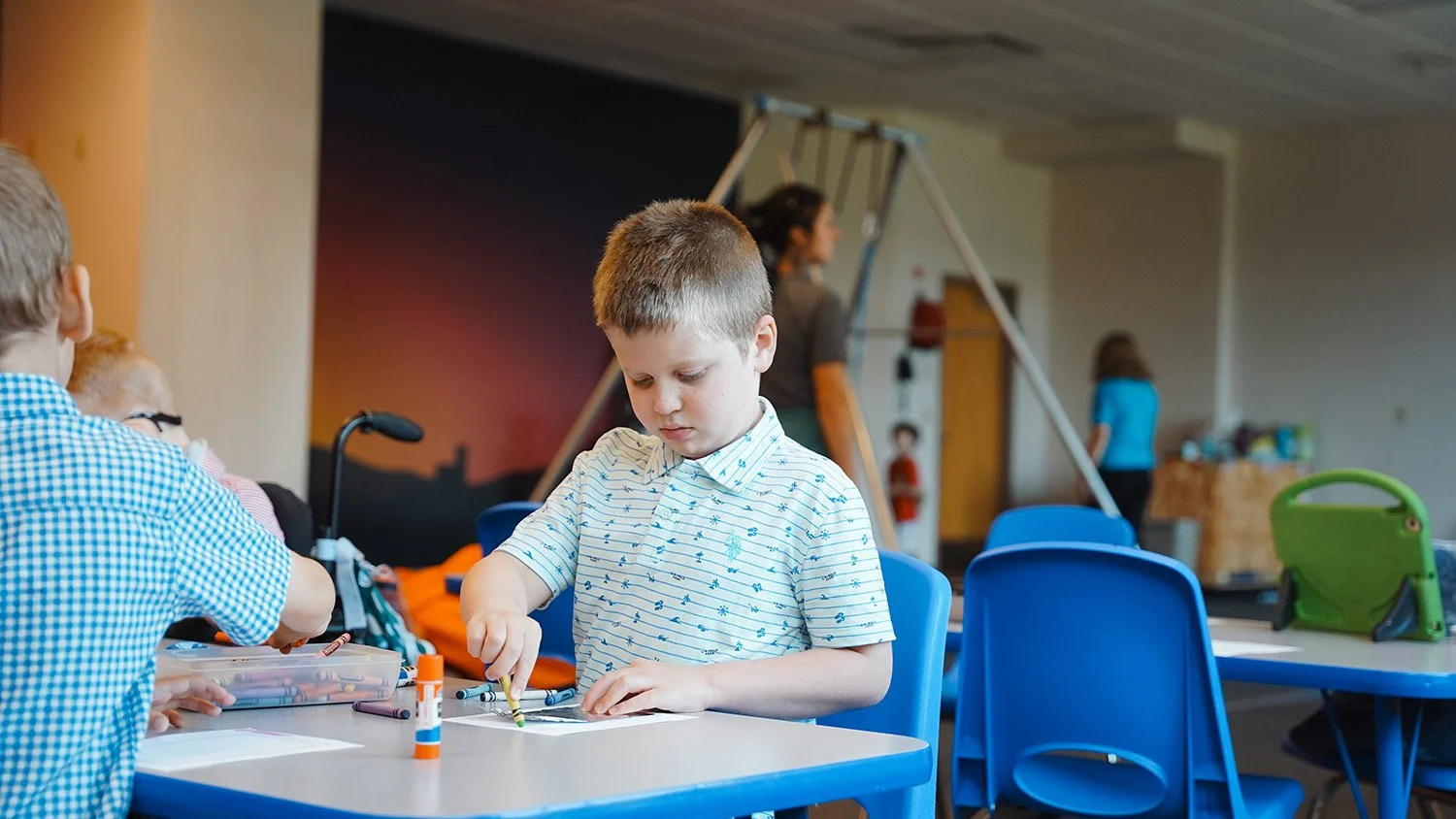 A young boy with short brown hair wearing a white shirt with blue patterns, focused on a craft project at a classroom table. Other children and adults are in the background, with some children playing and others sitting at tables in a room with a blue wall and playground equipment.