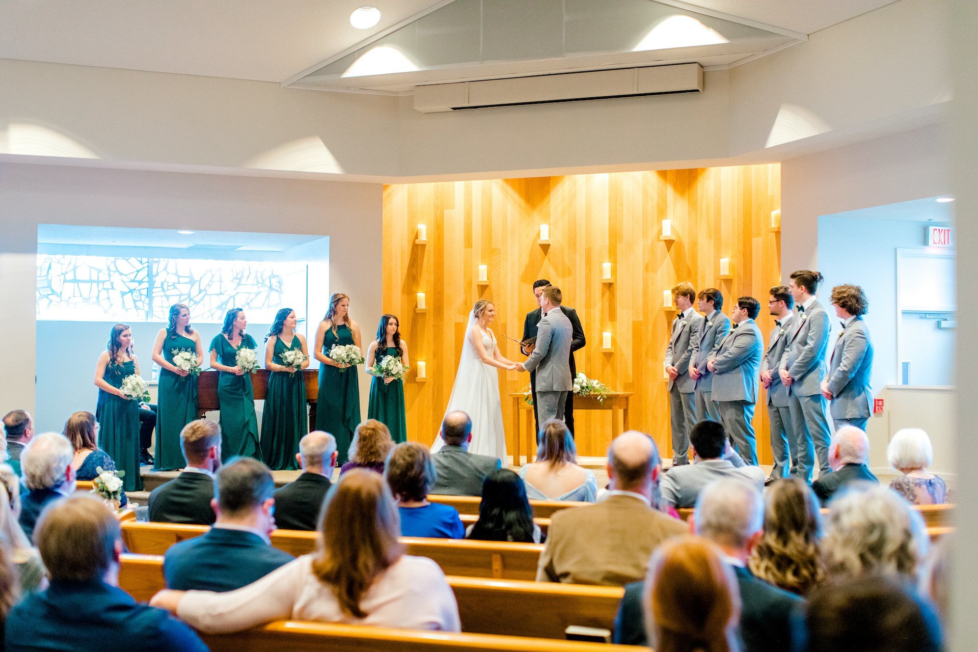 A wedding ceremony with a bride and groom holding hands at the altar, surrounded by bridesmaids in green dresses and groomsmen in gray suits. Guests are seated, watching the ceremony in a church setting with wood paneling and soft lighting.