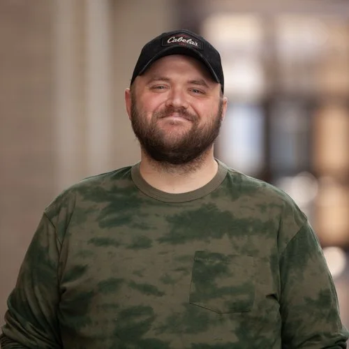 A smiling man wearing a Cabela's cap and a camouflage-patterned shirt, standing in a softly blurred indoor setting.
