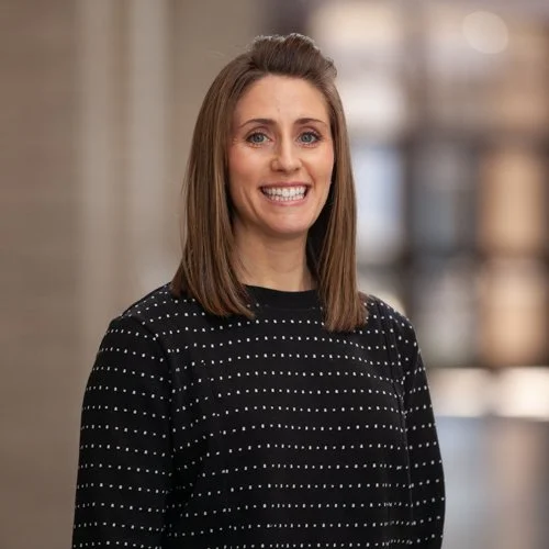 Woman smiling in a dark polka dot sweater, standing indoors with blurred background.