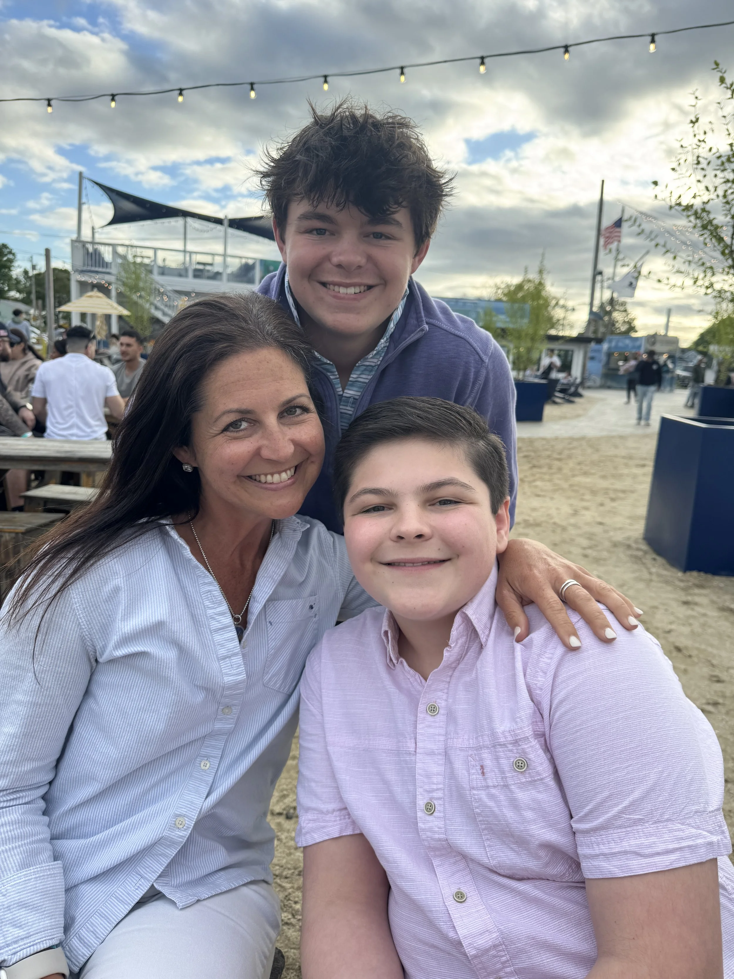 A woman and two boys smiling outdoors at a park or festival with string lights, sand, and flags in the background.