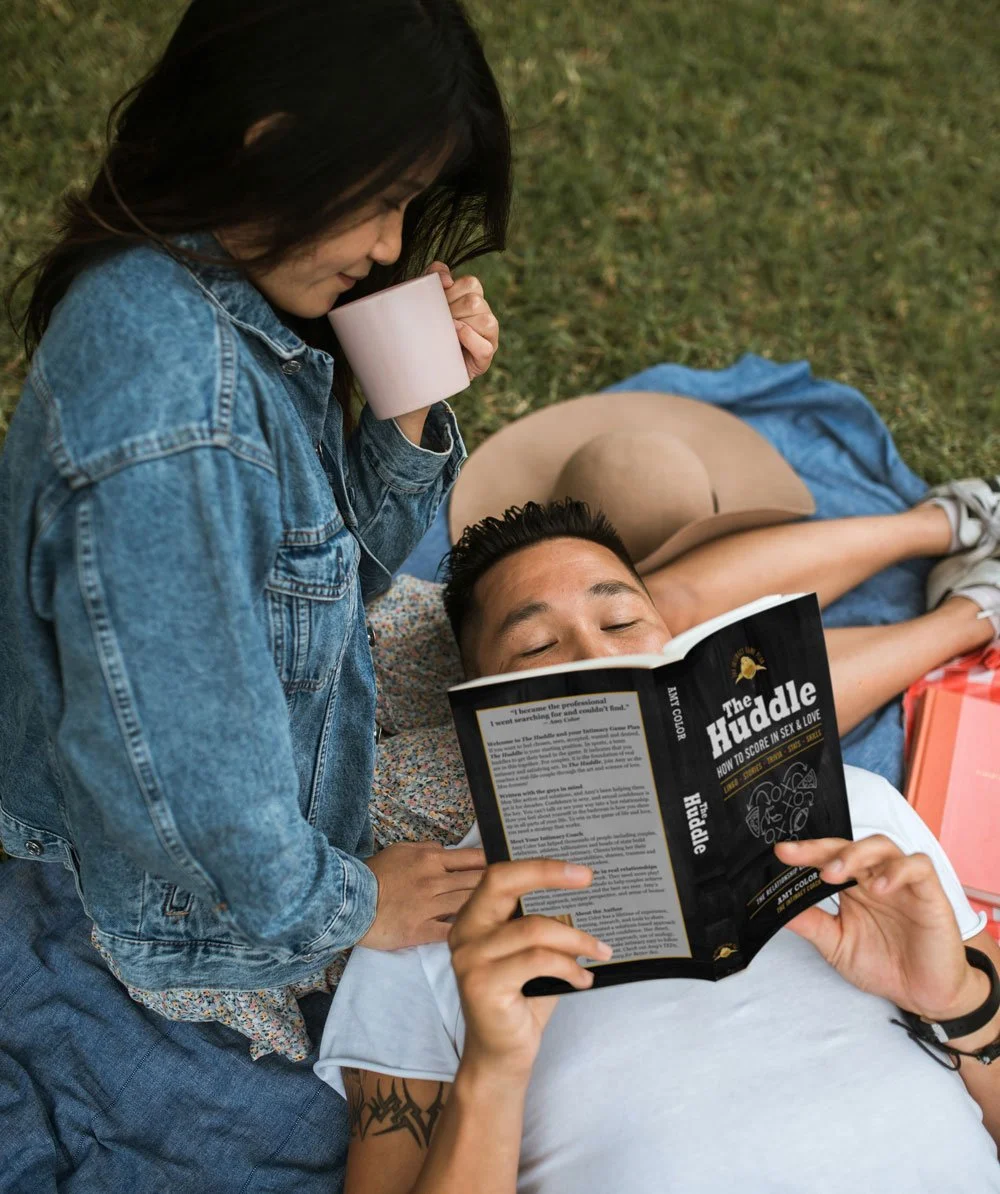 A couple relaxes on a picnic blanket outdoors. The man lies back reading “The Huddle: How to Score in Sex & Love” by Amy Color, while the woman stands beside him sipping from a pink mug, smiling. The moment feels cozy, connected, and curious.