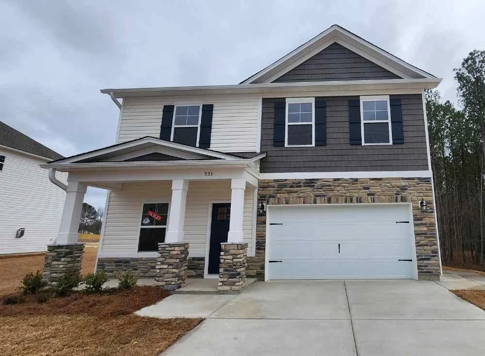 New two-story house with front porch, stone accents, paper 'sold' sign in window, attached garage, and well-maintained yard.