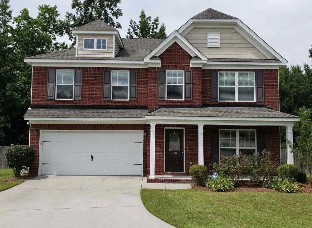 A two-story house with a brick exterior, gray roof, and white gutters. It has black shutters on the windows, a white garage door, and a front door with the number 390. There are small landscaped bushes and plants in the front yard with green grass.