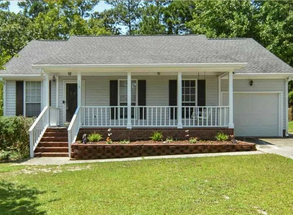 Front view of a house with a front porch, white railing, and a garage, surrounded by green lawn and trees.
