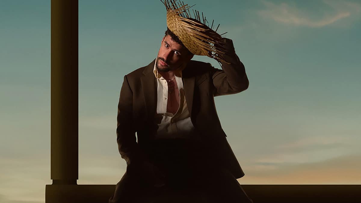 Bad Bunny holding his hat and sitting on top of the goal post.