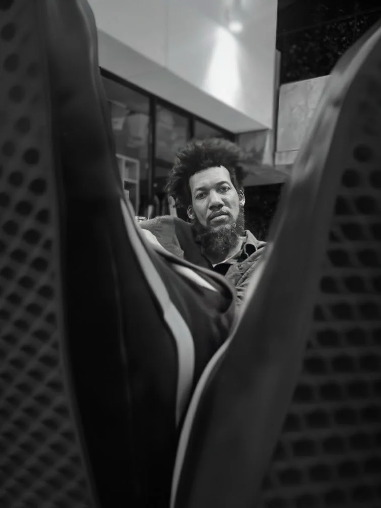 A black and white photo of a man with curly hair and a beard looking at the camera, taken from a low angle through the gap between two chairs.