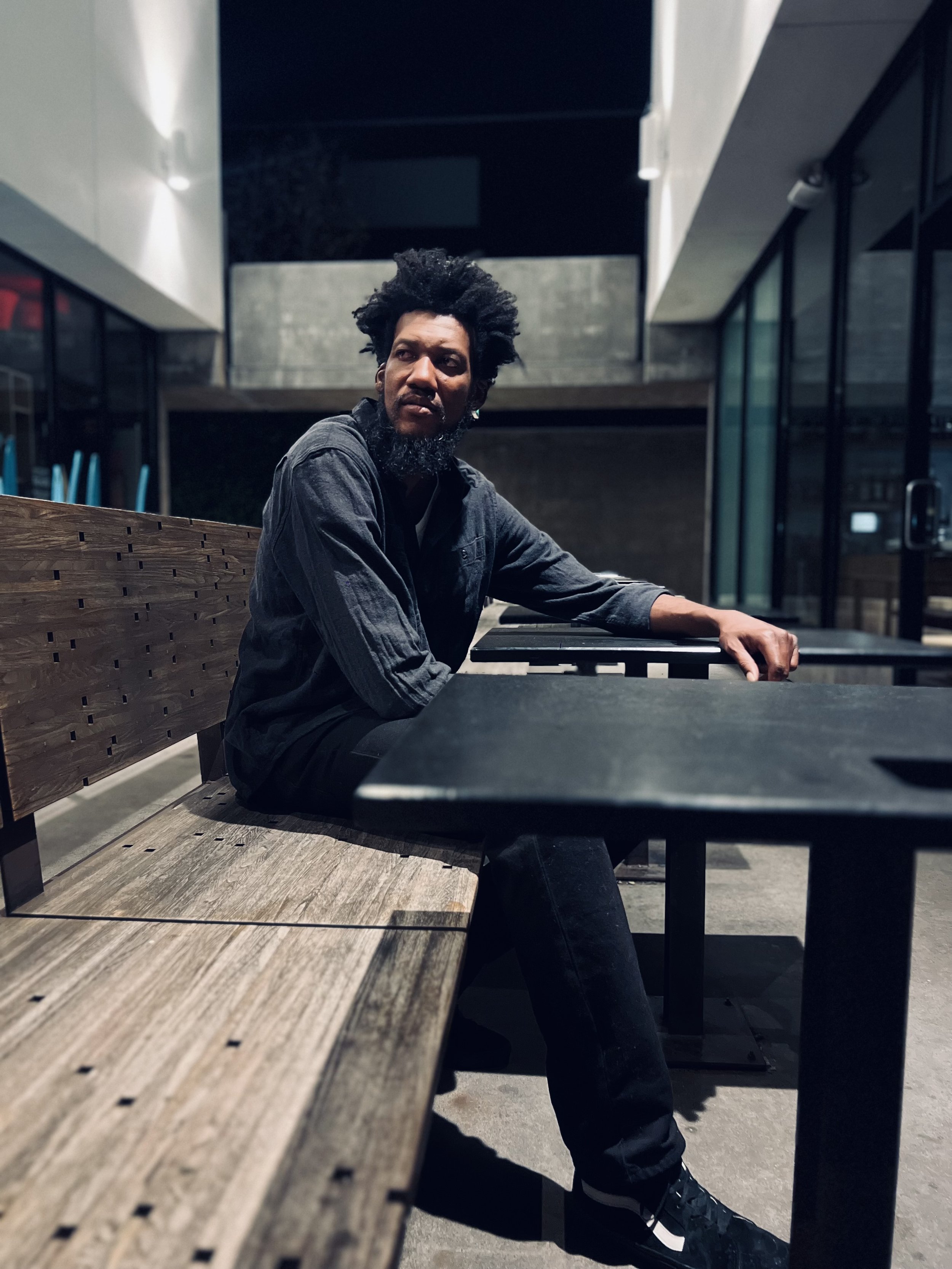 A man with an afro hairstyle and a beard sitting at an outdoor table at night in an urban setting, wearing a dark shirt and jeans.