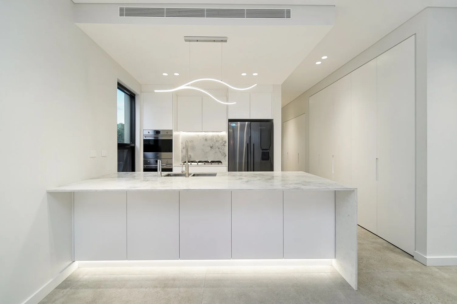 Modern, minimalist kitchen with white cabinets, marble countertops, stainless steel appliances, and a wave-shaped pendant light fixture.