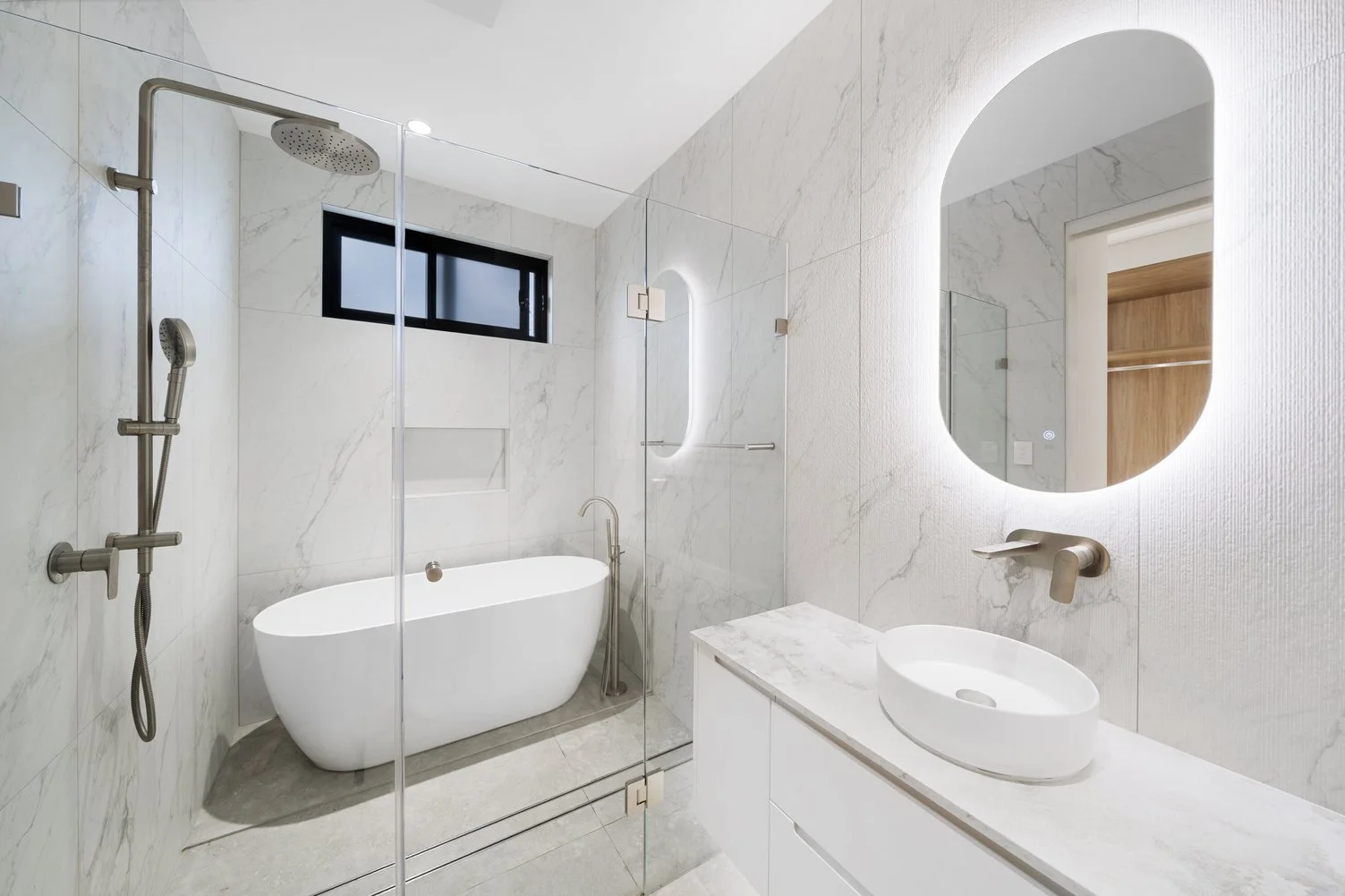 Modern bathroom with white marble walls, a freestanding bathtub, a glass shower enclosure, a round vessel sink on a marble countertop, a backlit mirror, and a small window.