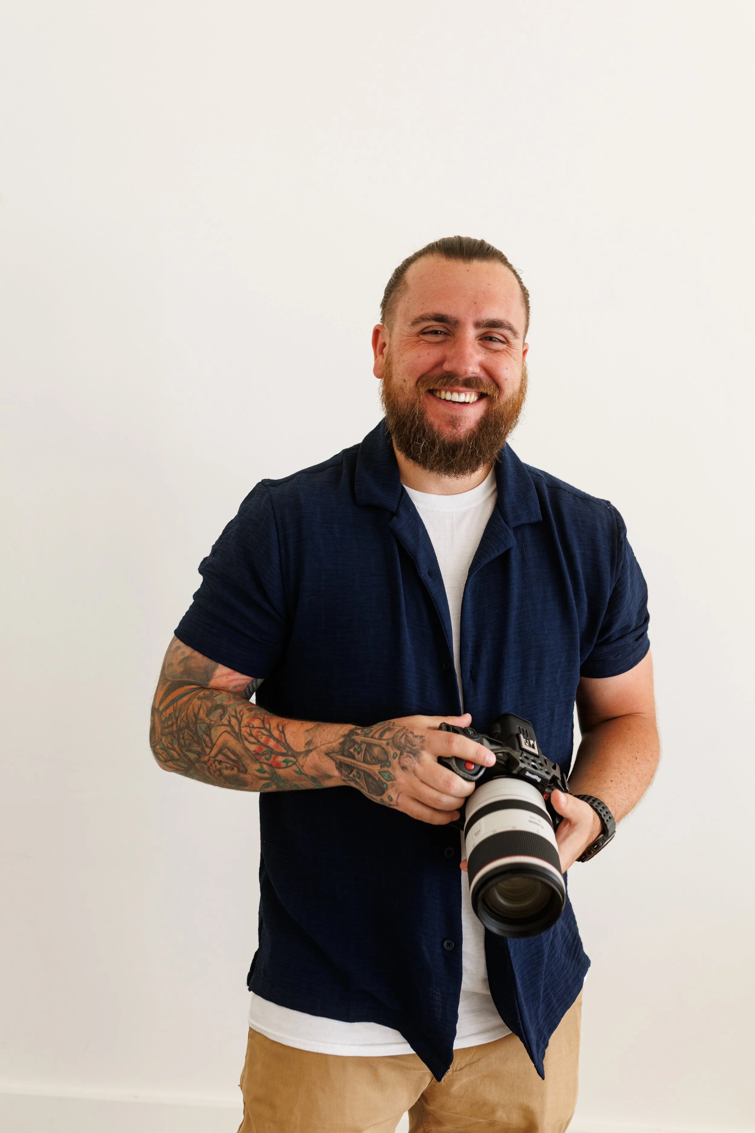 A man with a beard and tattoos holding a professional camera with a large zoom lens, standing against a plain white wall.