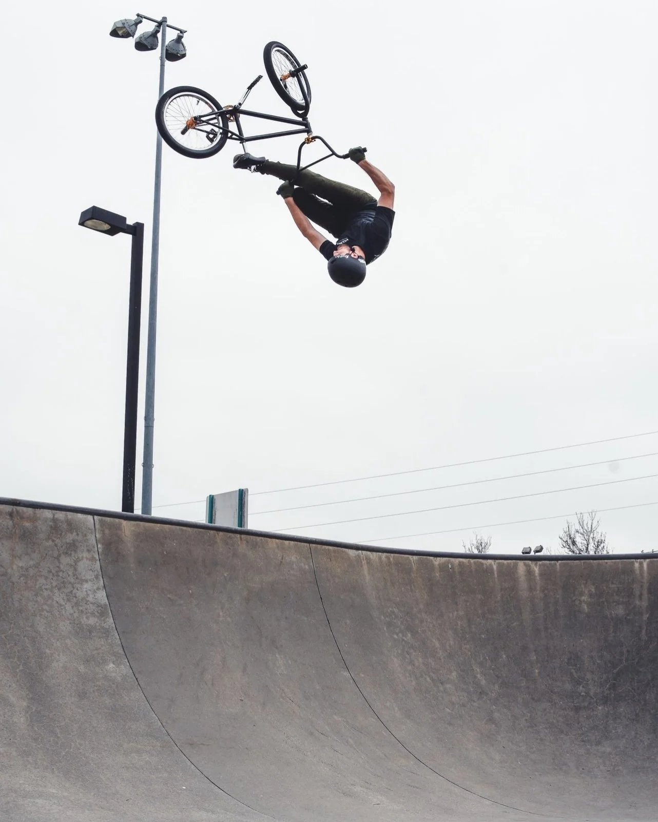 A person performing a BMX bike stunt in a skate park, captured mid-air upside down above a concrete ramp, wearing a helmet and black clothing.