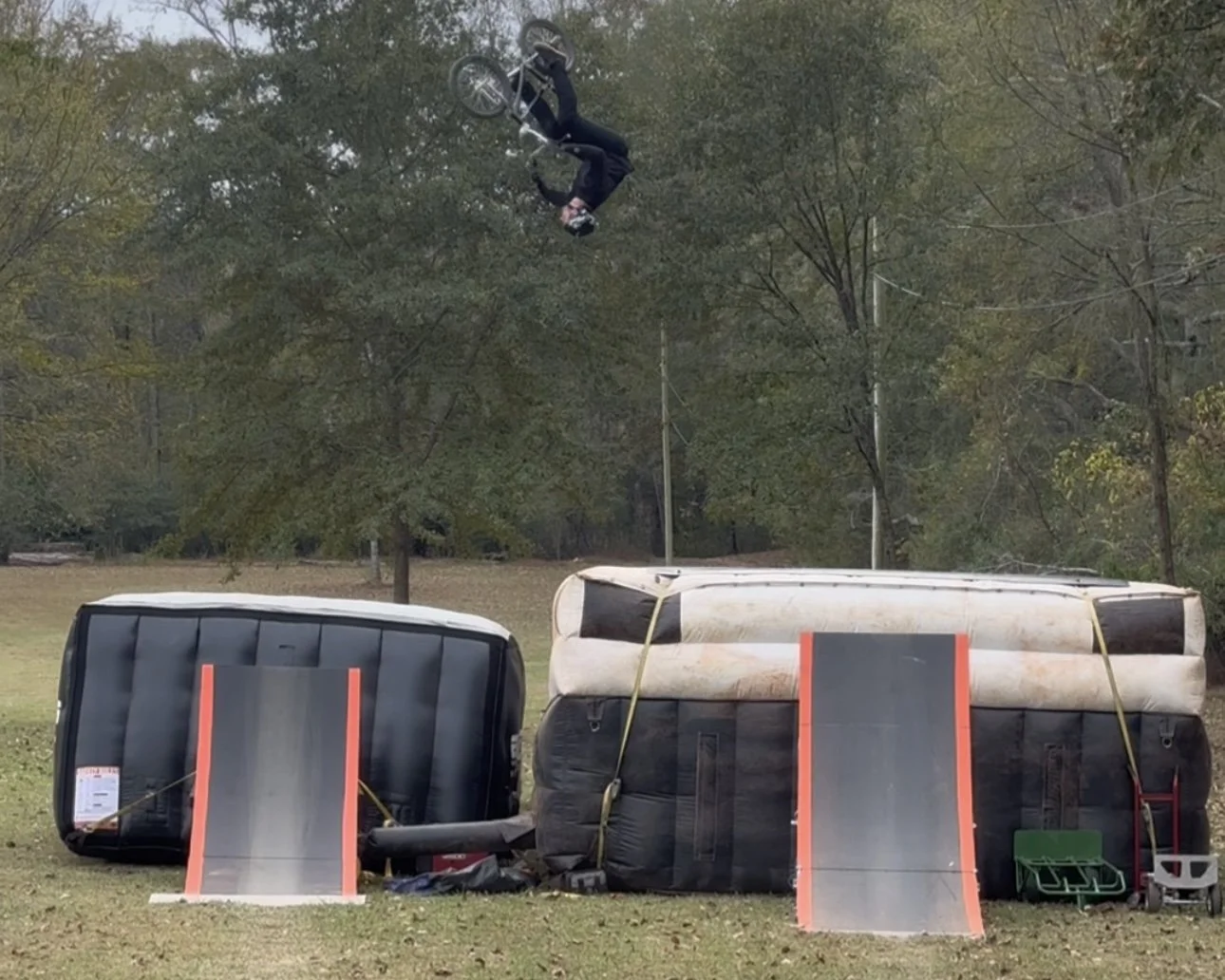 A person performing a BMX bicycle stunt above two inflatable ramps, set in an outdoor park area with trees.