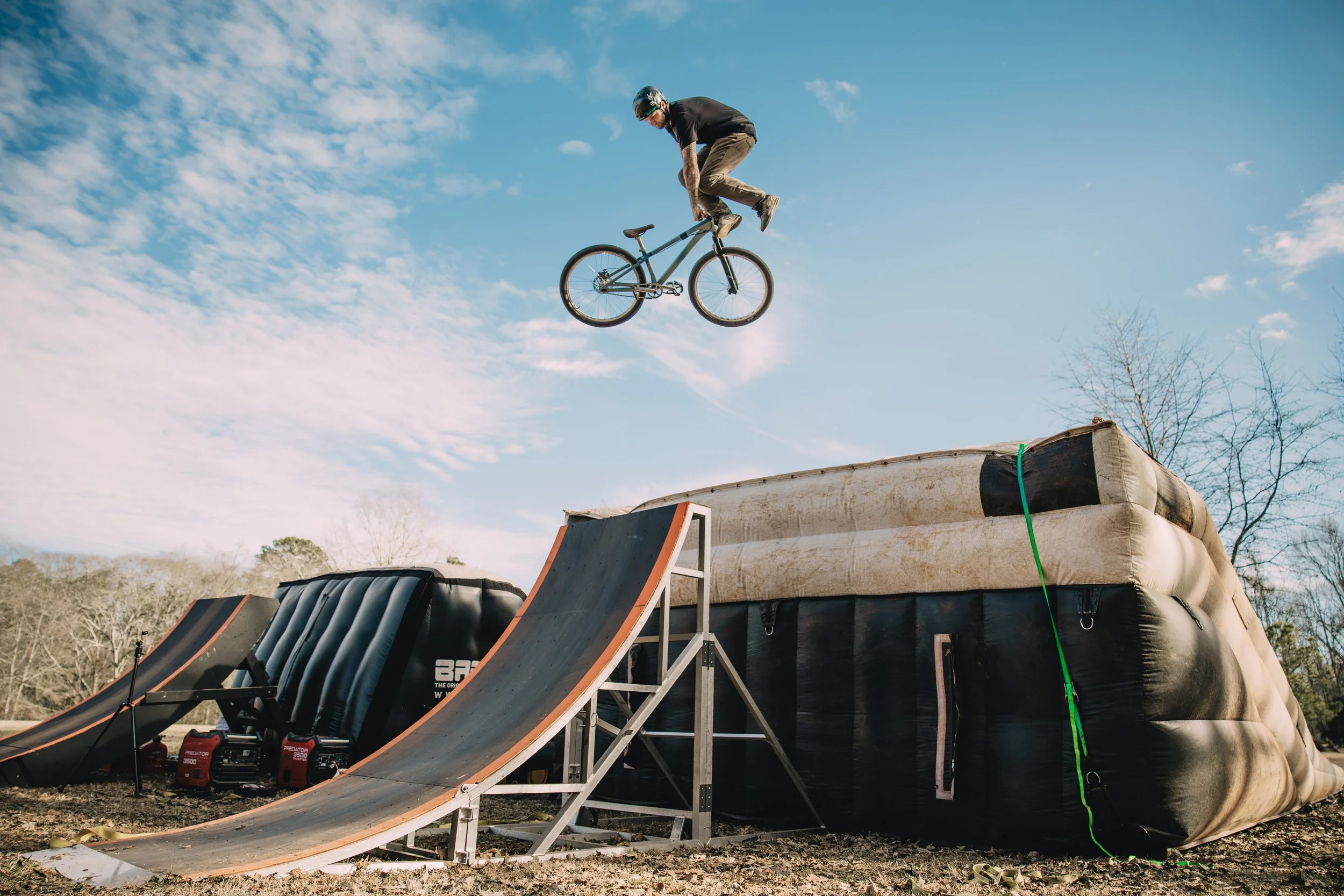 A person performing a BMX bicycle stunt above two inflatable ramps, set in an outdoor park area with trees.