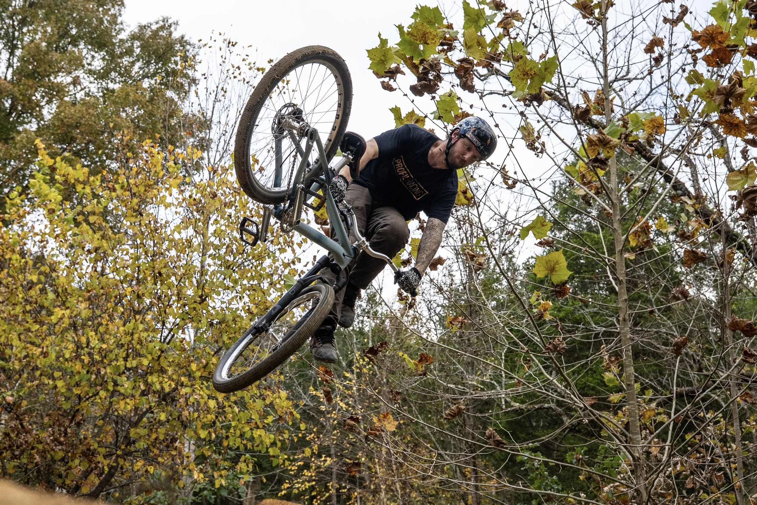 A man performing a BMX stunt on a dirt track in a wooded area during fall, with trees and leaves around him.