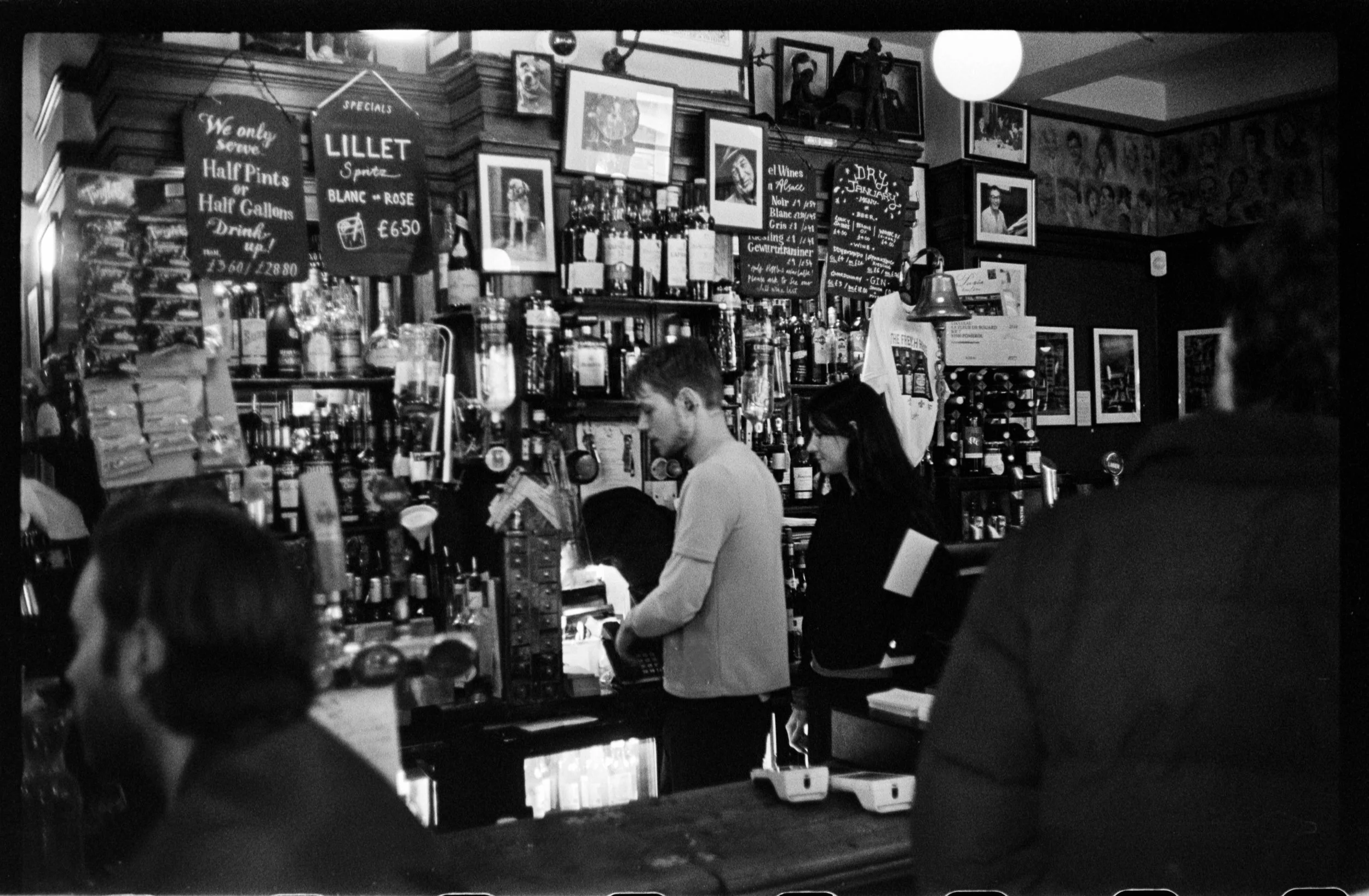 Black and white photo of people standing at a bar counter inside a pub, with shelves of bottles and artwork on the walls.