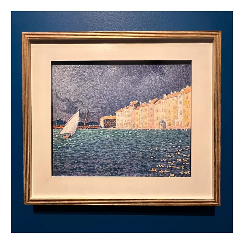 Painting of a sailboat on water near a row of buildings along the shoreline under a dark cloudy sky, framed and hung on a blue wall.