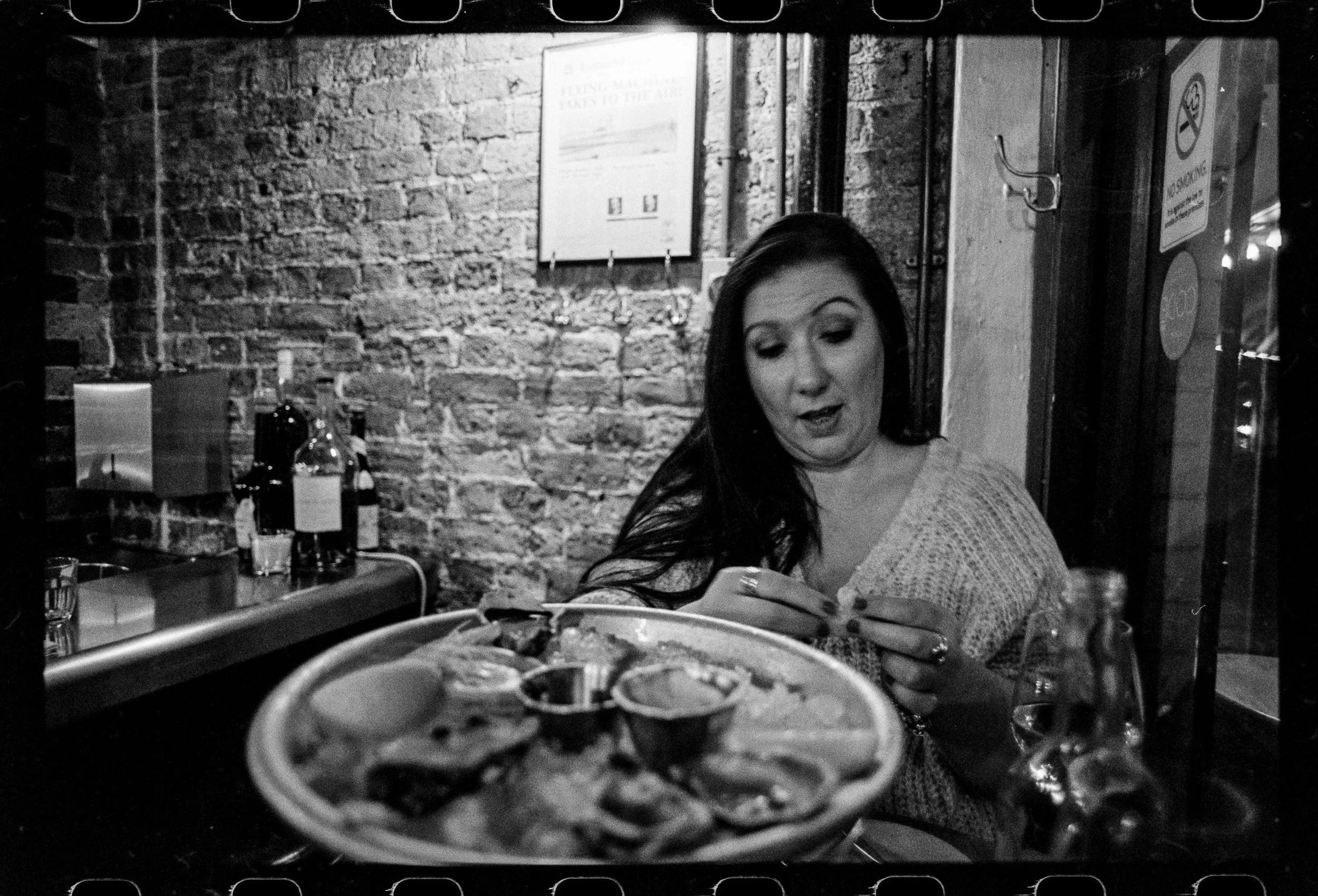 A woman with long dark hair sitting at a restaurant table, looking at her phone with a smile. The background features a brick wall, a framed picture, and bottles on the table, with a pizza in the foreground.