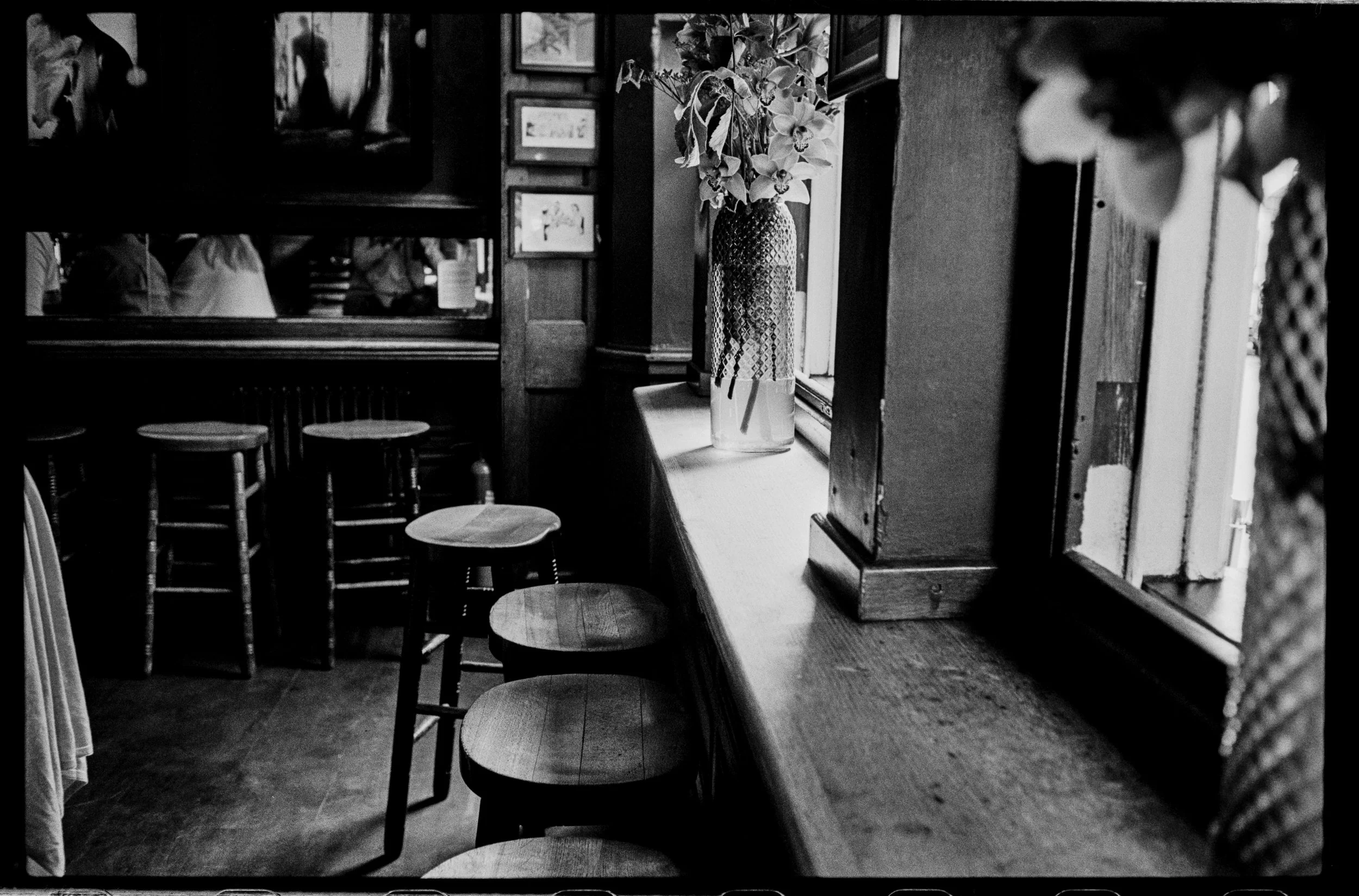 Black and white photo of a cozy corner in a restaurant or cafe featuring a window with a vase of flowers on the windowsill, several stools, and a wall with picture frames.