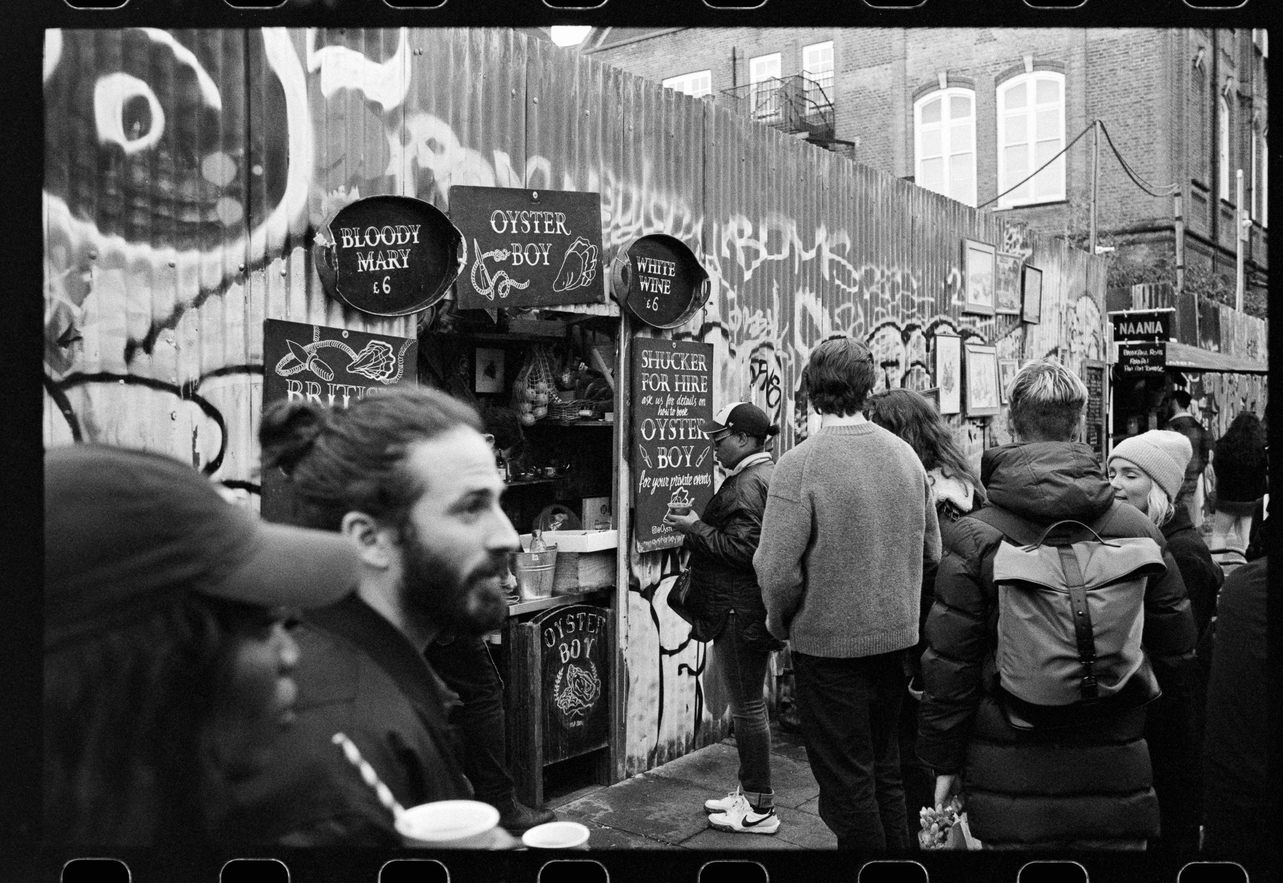 A black and white photo taken at an outdoor market or fair. Several people are seen from behind, standing in front of a stall decorated with chalkboard signs. The signs advertise seafood items, such as "Bloody Mary", "Oyster Boy", and "White Wine", w