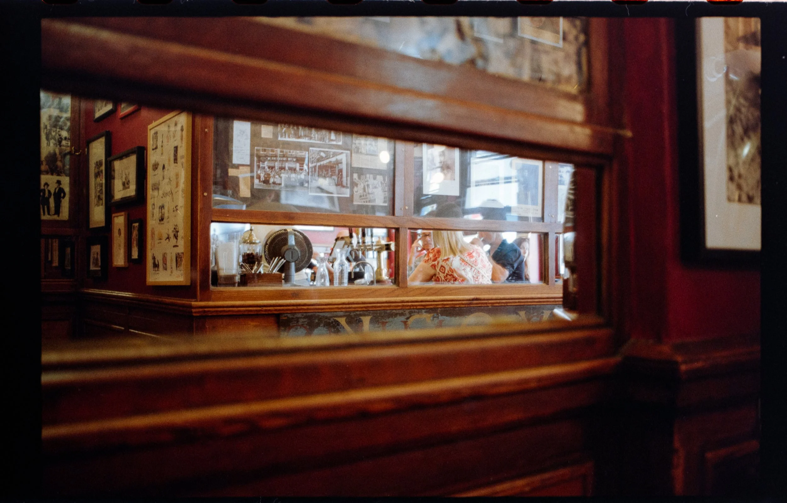View of a restaurant interior through a wooden framed window. Visible are several framed pictures and a woman wearing an orange patterned top sitting at a table with drinks.