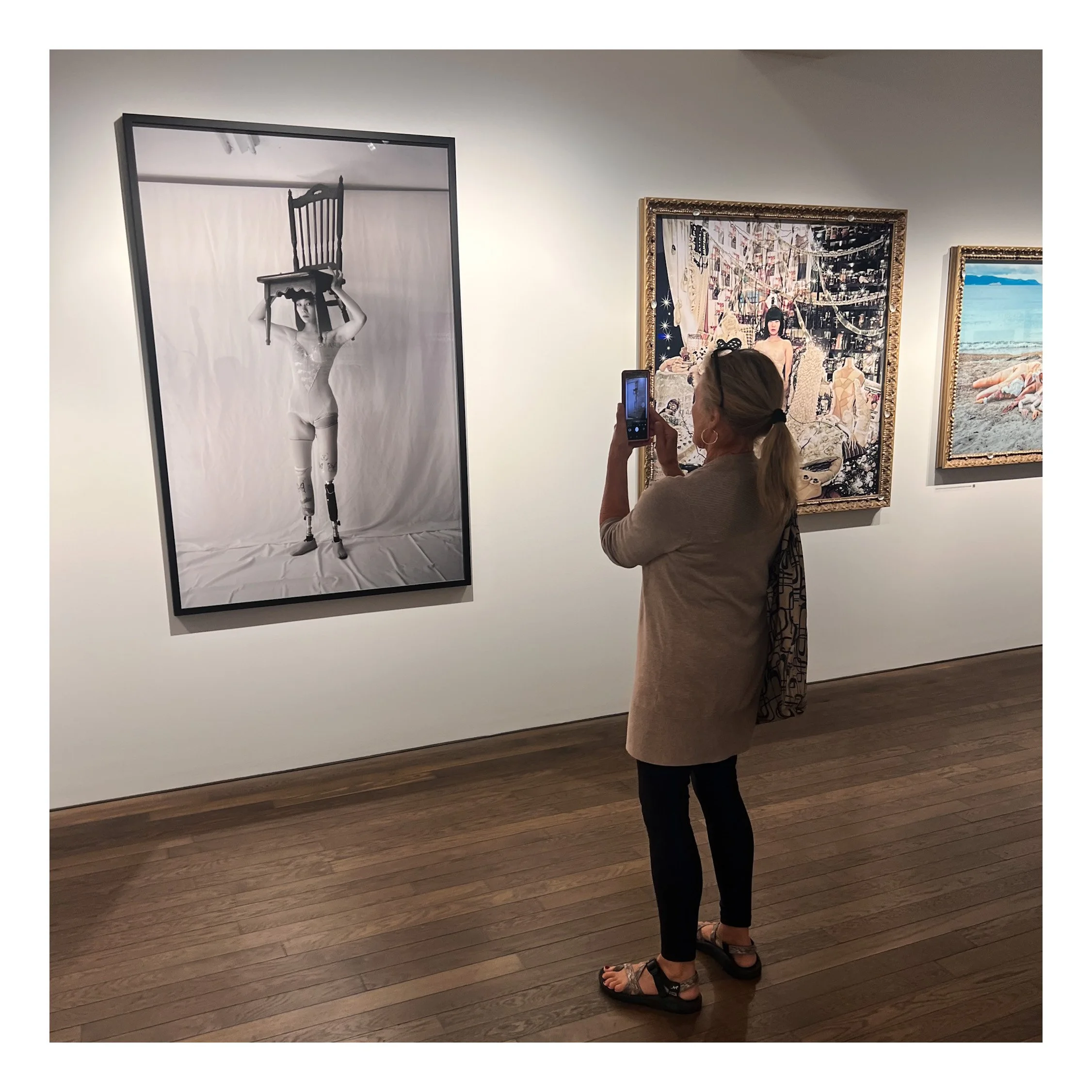 A woman with brown hair tied back taking a photo of artwork with her smartphone in a gallery. The artwork on the left is a black and white photograph of a woman holding a chair above her head, wearing lingerie and tights. The walls are white, and the