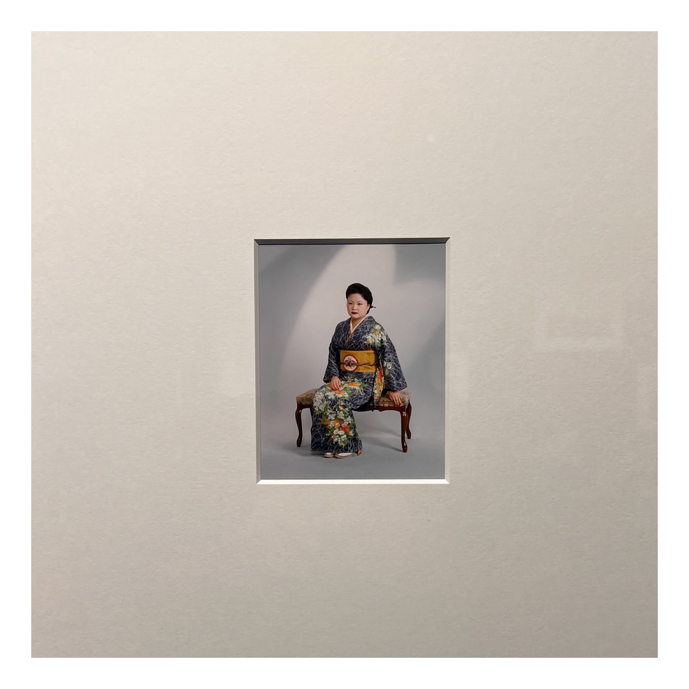 A woman dressed in traditional Japanese kimono standing in a gallery with a white wall and an abstract portrait painting behind her.