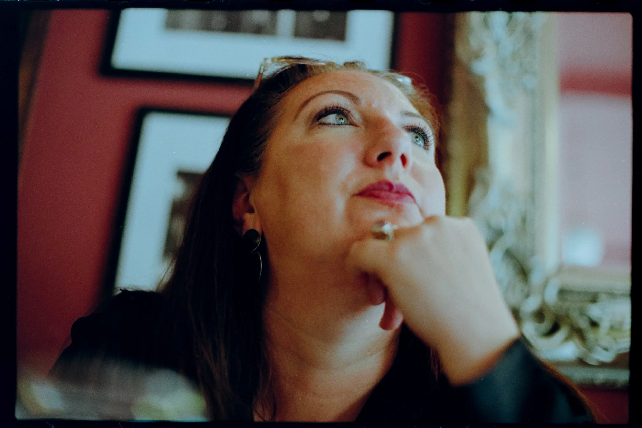Close-up of a woman with reddish-brown hair, looking up and slightly to the right, with a thoughtful expression. She is indoors, with framed pictures on a red wall behind her and a window with curtains to her right.