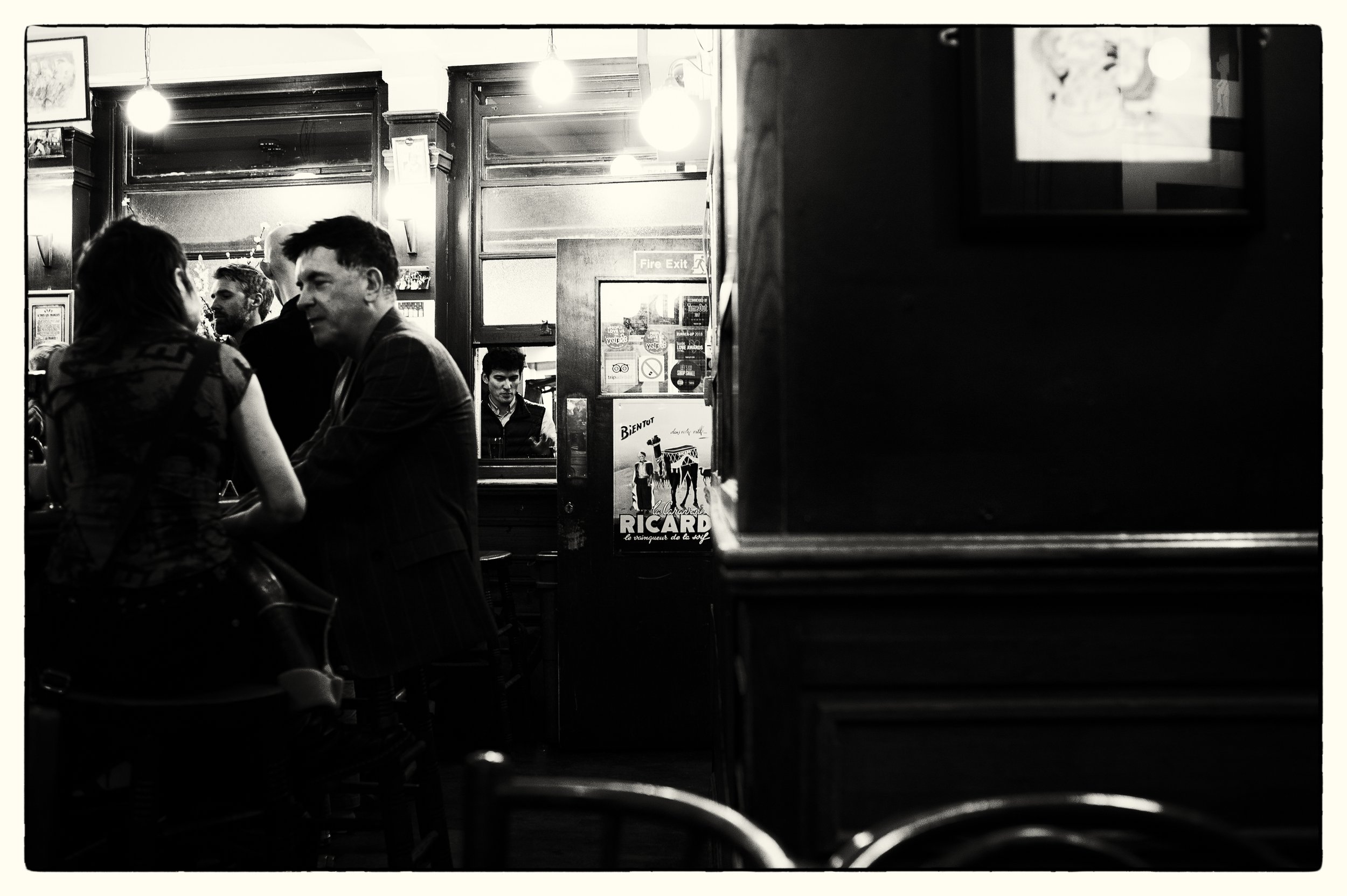 Black-and-white photo of a busy pub or bar with people talking. The foreground shows part of a chair and a small table. The background features wooden walls, a mirror, posters, and a man seated behind the bar. Two hanging lights illuminate the scene.