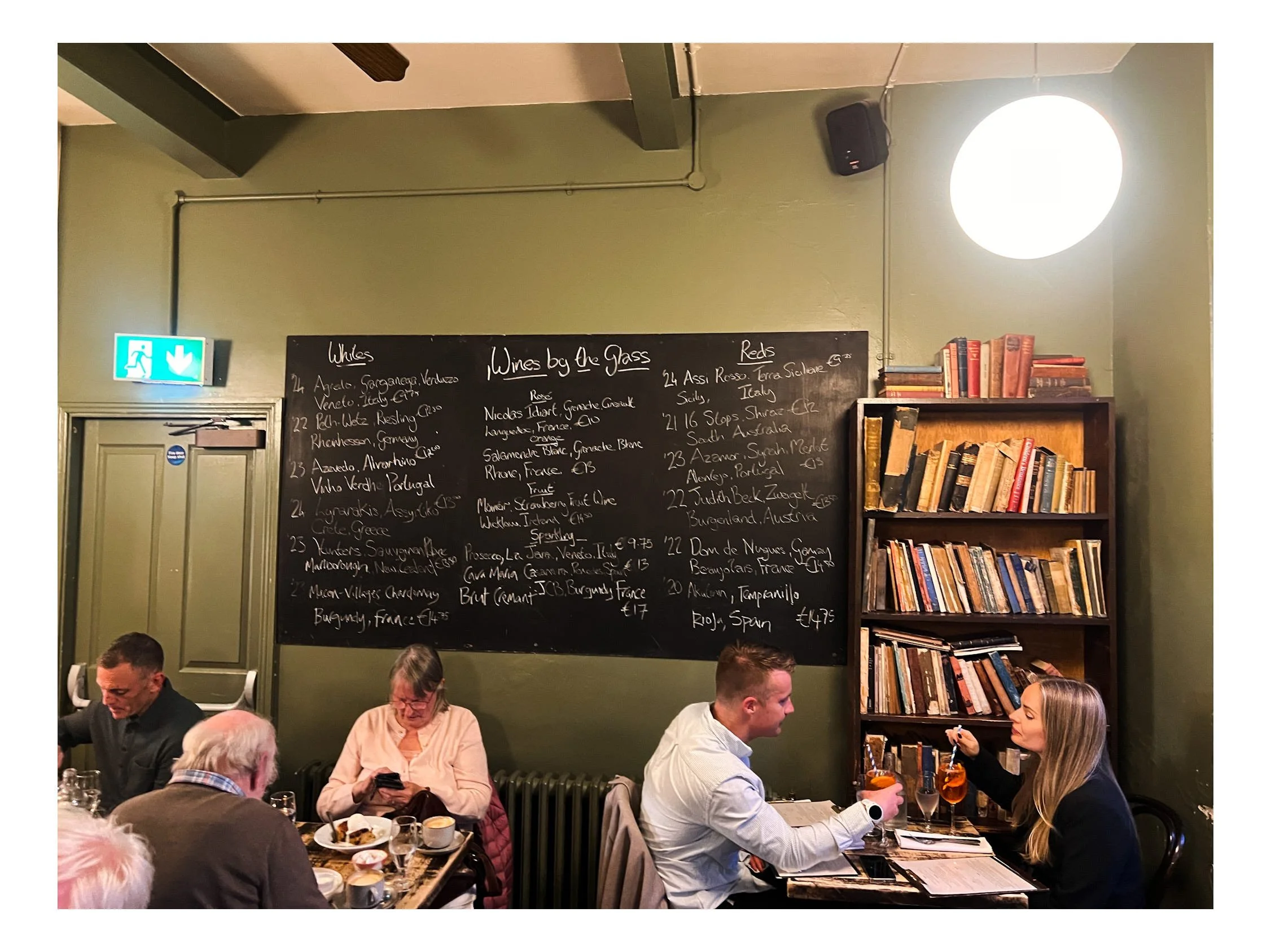 People dining and socializing in a restaurant with a chalkboard menu listing wines, a bookshelf, and an emergency exit sign.